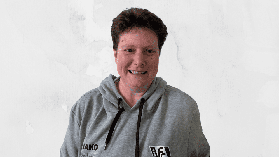 L&auml;chelnde Person in grauem Hoodie vor hellen, strukturierten Hintergrund.