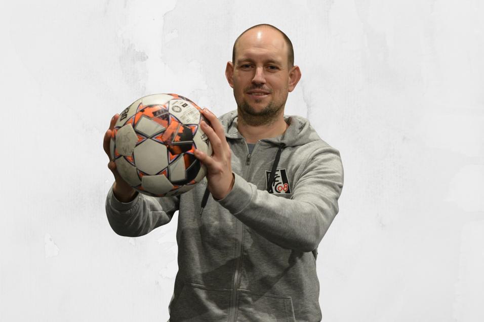 Mann h&auml;lt einen Fu&szlig;ball in beiden H&auml;nden, tr&auml;gt einen grauen Hoodie und steht vor einer hellen Hintergrundfl&auml;che.