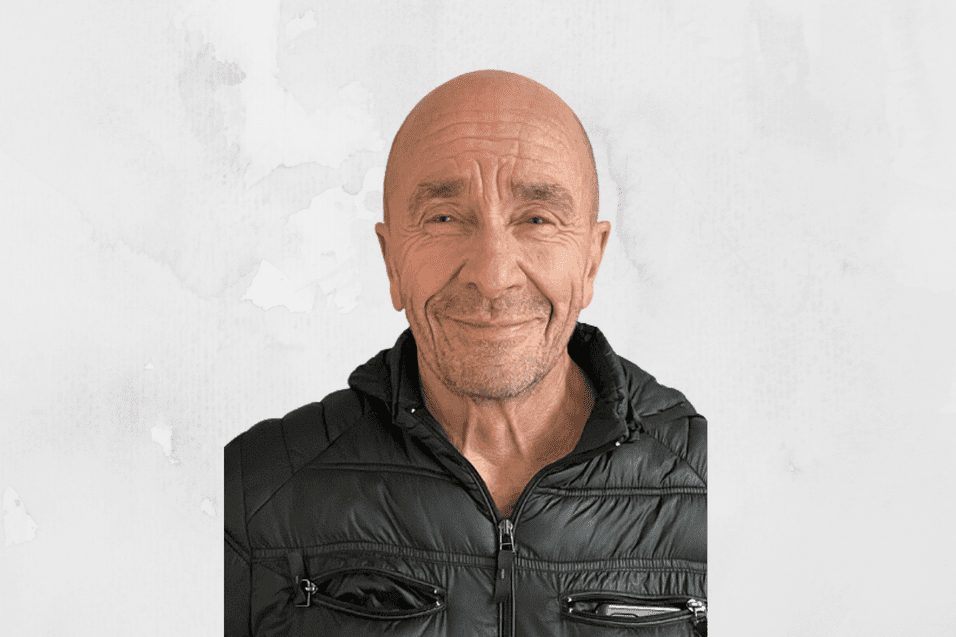 Portrait eines l&auml;chelnden Mannes mit Glatze, der eine schwarze Jacke tr&auml;gt, vor hellem, einfarbigem Hintergrund.