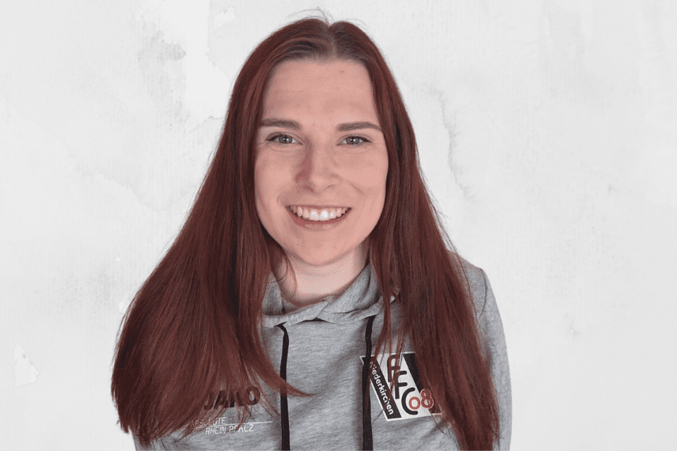 L&auml;chelnde Frau mit langen, roten Haaren, tr&auml;gt einen grauen Hoodie vor hellem, einfarbigen Hintergrund.