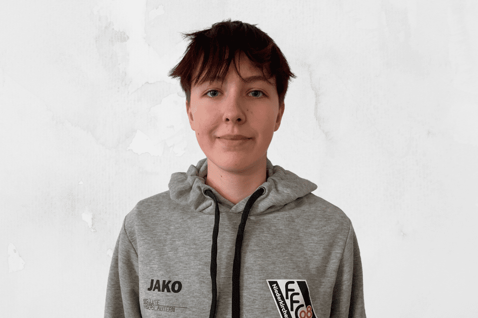 Portr&auml;t einer Person mit kurzen, braunen Haaren, die einen grauen Hoodie mit Logos tr&auml;gt, vor einer hellen, neutralen Wand.