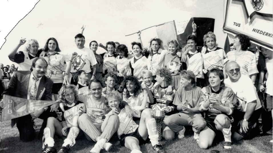 Gruppe von Fu&szlig;ballspielern und Supportern posiert freudig auf einem Platz, umgeben von Bannern und Pokalen.