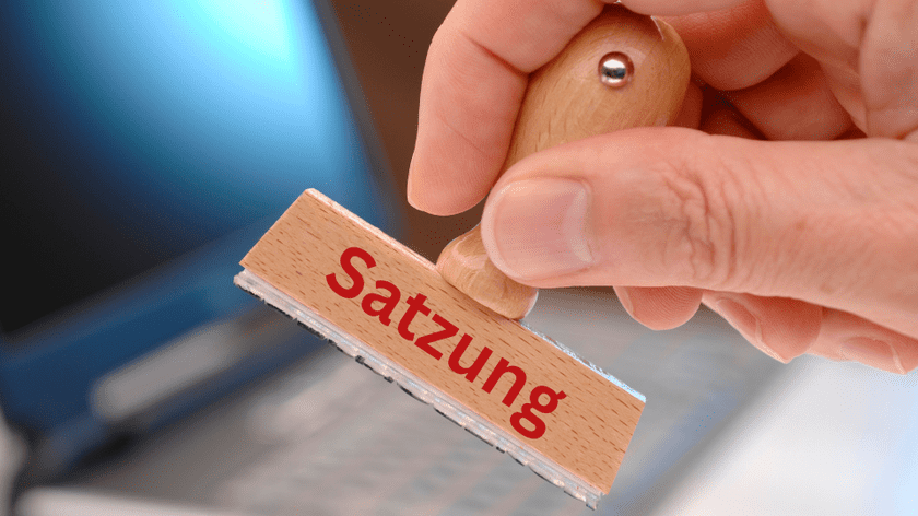 Hand h&auml;lt einen Holzstempel mit der Aufschrift &bdquo;Satzung&ldquo; &uuml;ber einem Laptop.
