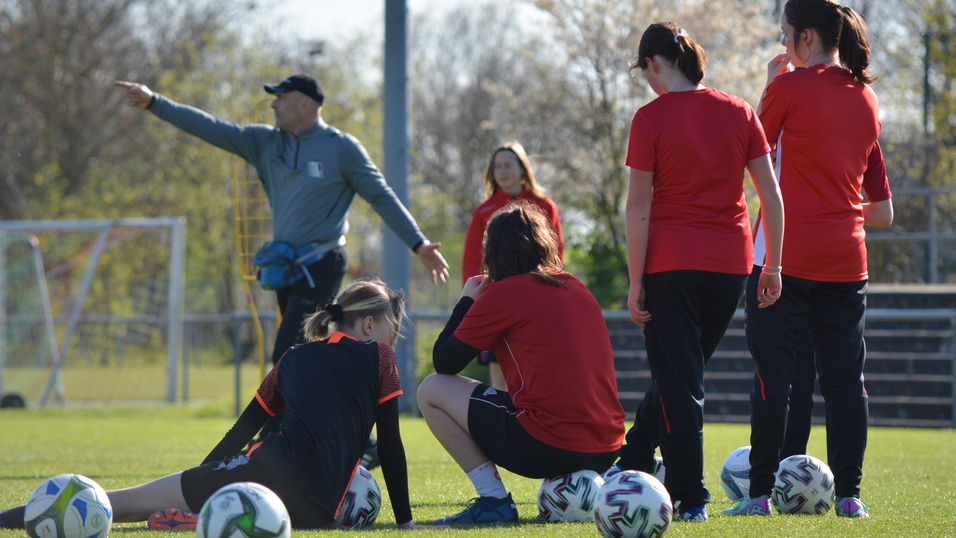 Trainingssituation auf einem Fu&szlig;ballplatz mit einer Trainerin und f&uuml;nf Spielerinnen in roten Trikots, einige sitzen, andere stehen.