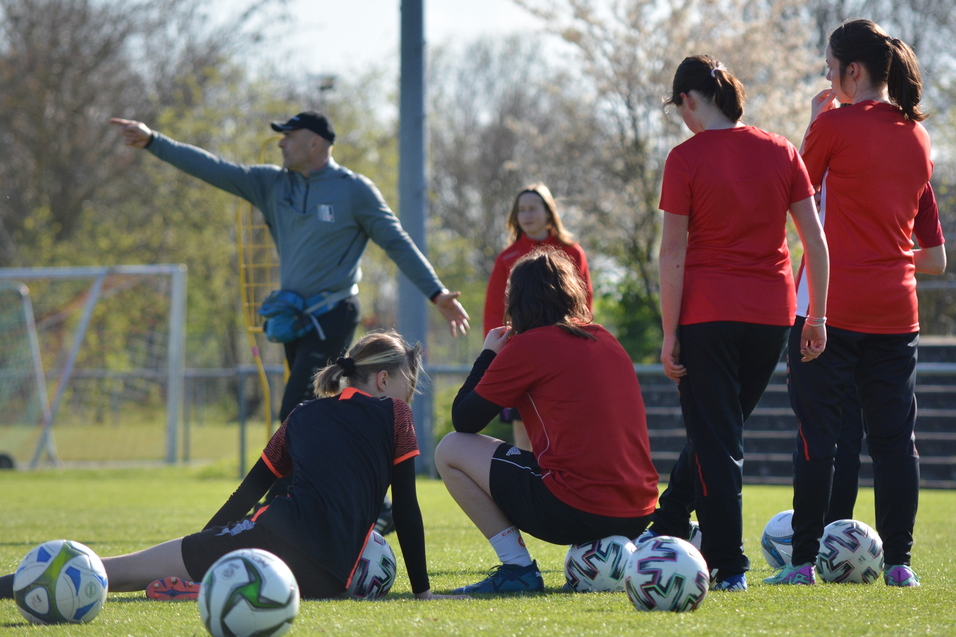 Trainingssituation auf einem Fu&szlig;ballplatz mit einer Trainerin und f&uuml;nf Spielerinnen in roten Trikots, einige sitzen, andere stehen.