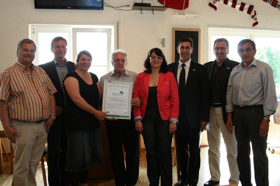 Gruppe von sieben Personen steht in einem Raum, einige halten ein Zertifikat. Wand mit Trikots dekoriert.