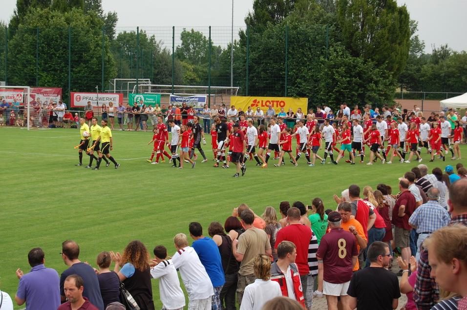Fu&szlig;ballmannschaften und Schiedsrichter betreten das Spielfeld, umgeben von jubelnden Zuschauern auf der Trib&uuml;ne.