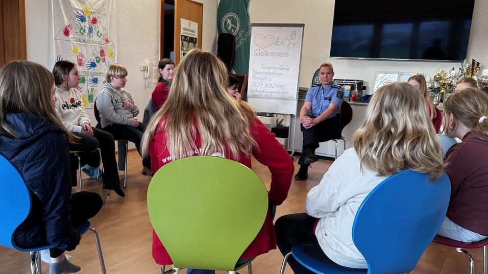 Gruppe von Kindern und Jugendlichen sitzt auf bunten St&uuml;hlen, w&auml;hrend eine Person im Vordergrund spricht.