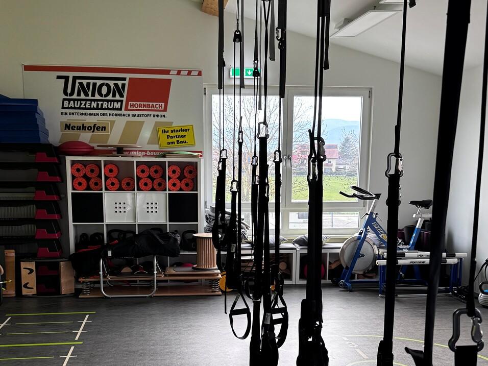 Nahaufnahme eines Fitnessraums mit mehreren Trainingsger&auml;ten und TRX-B&auml;ndern an der Wand, darunter schwarzer Boden.