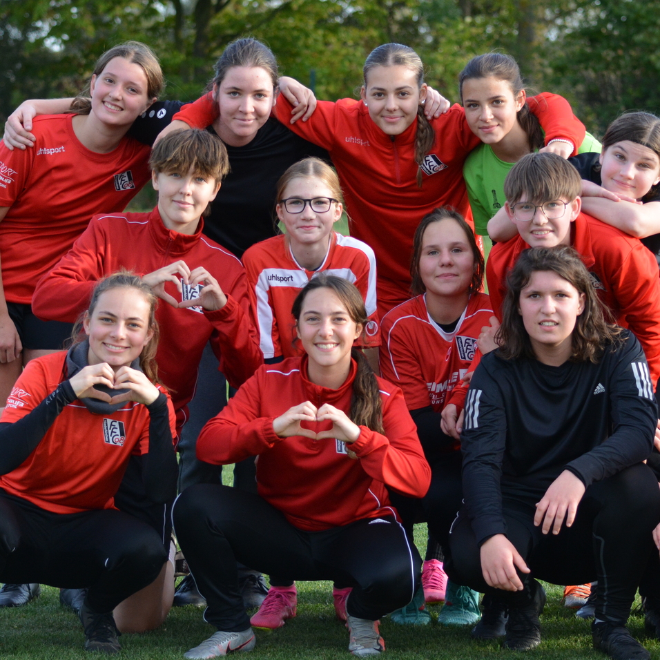 Gruppenausschnitt von 14 jungen Fu&szlig;ballspielerinnen in roten Trikots, die auf einem Rasenfeld posieren und ein Herz formen.