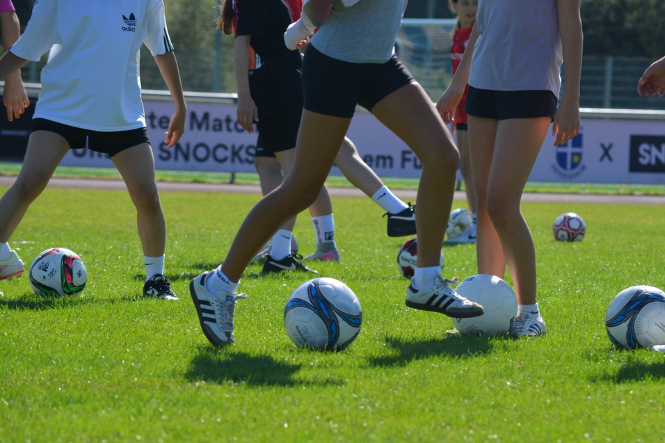 M&auml;dchen in Sportkleidung &uuml;ben im Stehen mit Fu&szlig;b&auml;llen auf einem gr&uuml;nen Rasenplatz.