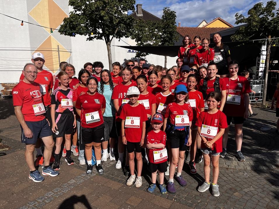 Gruppe von L&auml;ufern in roten Shirts versammelt sich vor einer Wand, bereit f&uuml;r ein Sportevent, mit Nummern auf ihren Shirts.