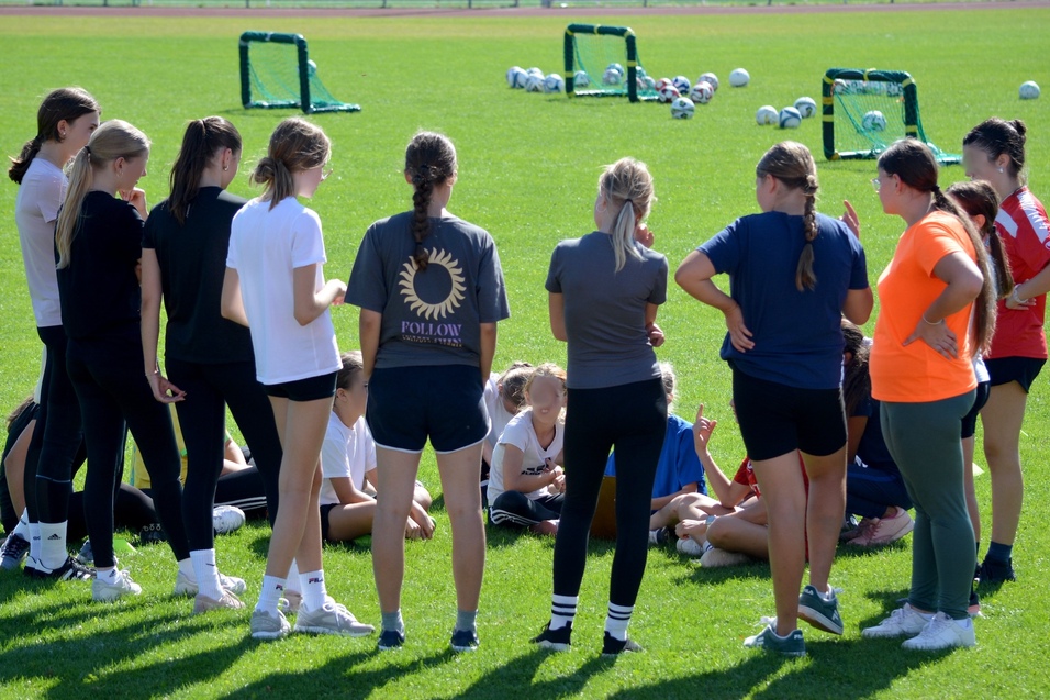 Gruppe junger Frauen steht um eine sitzende Gruppe auf einem Fu&szlig;ballfeld, im Hintergrund sind Tore und B&auml;lle zu sehen.