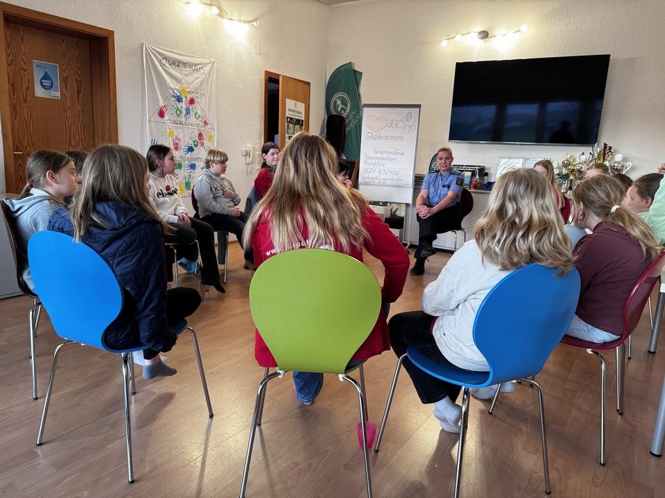 Gruppe von Kindern und Jugendlichen sitzt auf bunten St&uuml;hlen, w&auml;hrend eine Person im Vordergrund spricht.