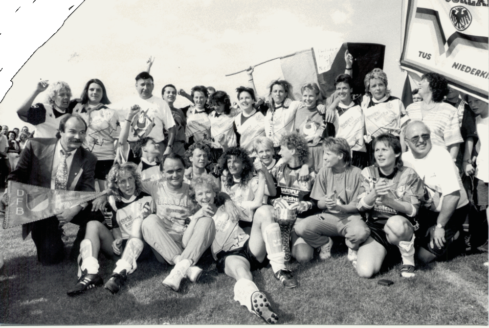 Gruppe von Fu&szlig;ballspielern und Supportern posiert freudig auf einem Platz, umgeben von Bannern und Pokalen.