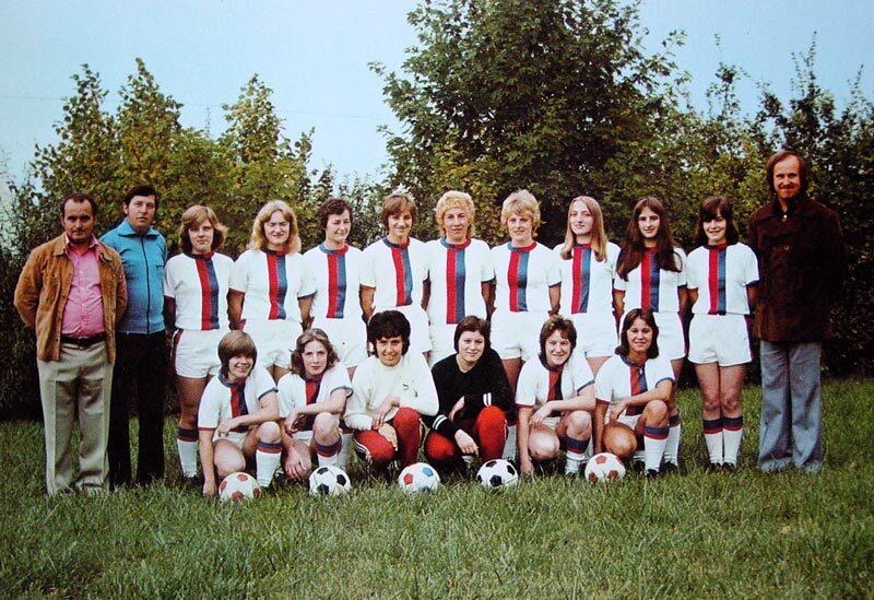 Gruppenfoto einer Frauenfu&szlig;ballmannschaft mit Trainer, in wei&szlig;en Trikots mit roten und blauen Streifen, auf einer Wiese.