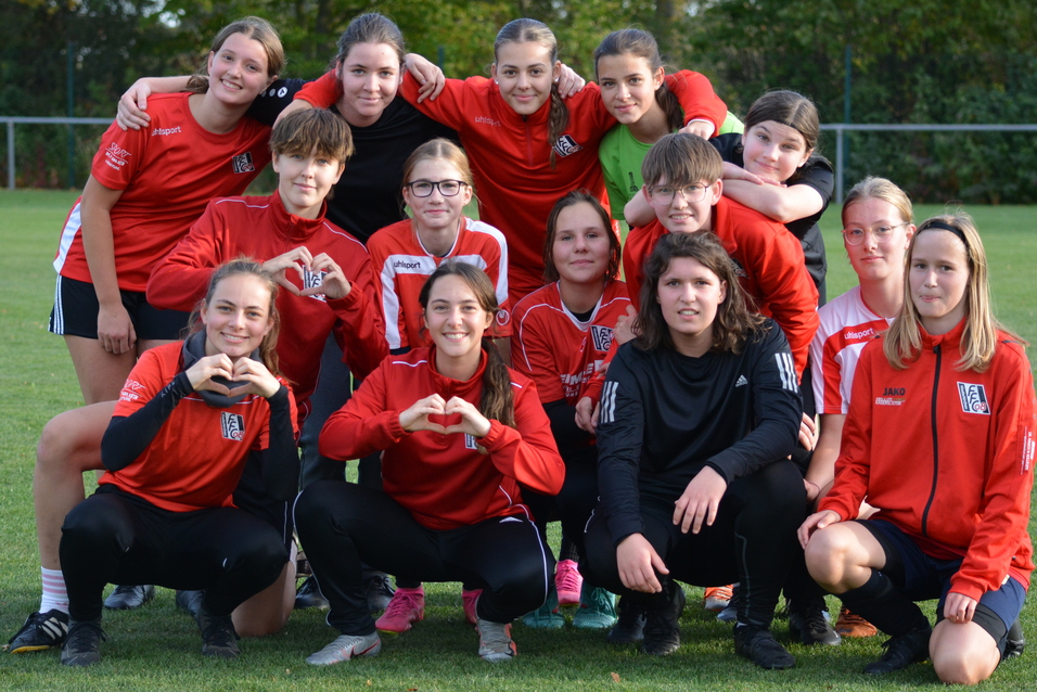 Gruppenausschnitt von 14 jungen Fu&szlig;ballspielerinnen in roten Trikots, die auf einem Rasenfeld posieren und ein Herz formen.