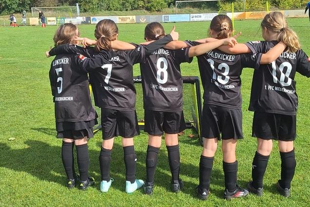 Fünf Mädchen in schwarzen Fußballtrikots stehen mit dem Rücken zur Kamera auf einem Rasenplatz.