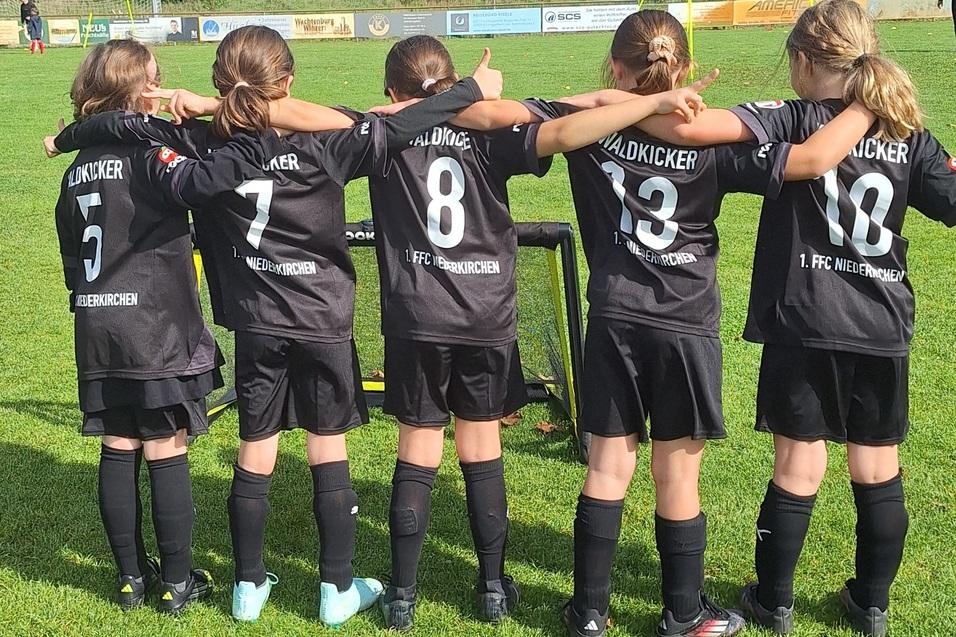 F&uuml;nf M&auml;dchen in schwarzen Fu&szlig;balltrikots stehen mit dem R&uuml;cken zur Kamera auf einem Rasenplatz.