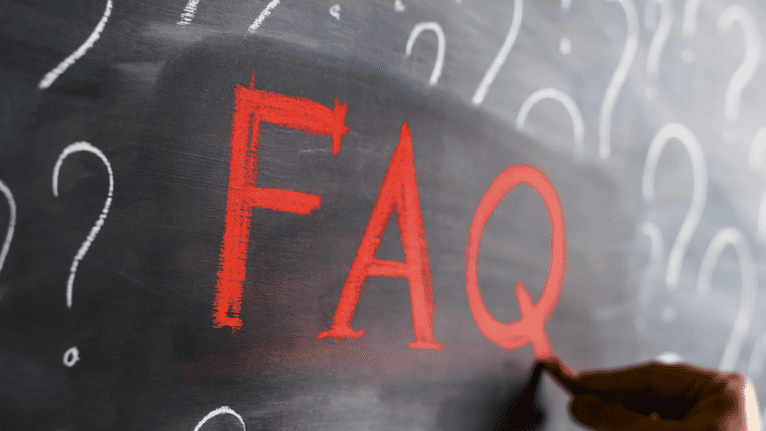 Nahaufnahme von einer Hand, die mit roter Kreide "FAQ" auf eine schwarze Tafel mit wei&szlig;en Fragezeichen schreibt.