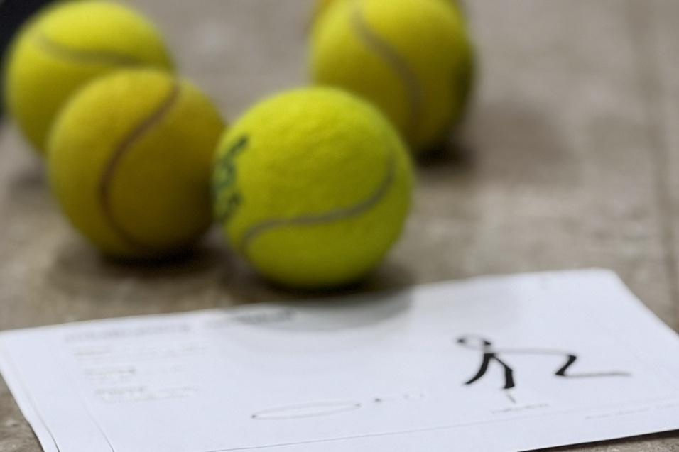 Vier gelbe Tennisb&auml;lle liegen auf einem Sportboden neben einem Blatt Papier mit einer einfachen Zeichnung.