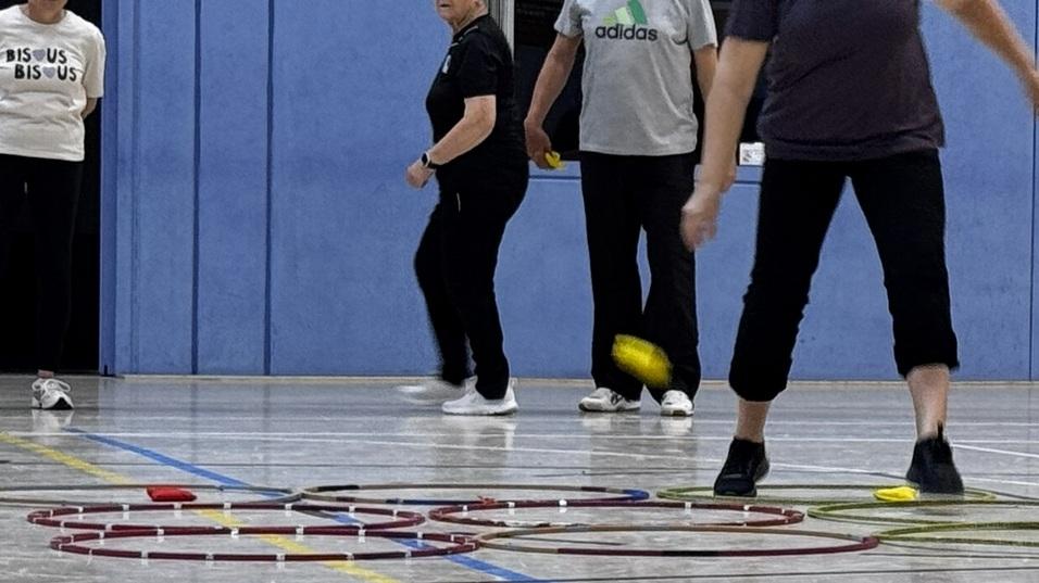 Eine Gruppe von Personen spielt Sport in einer Sporthalle, einige laufen, w&auml;hrend andere zuschauen.