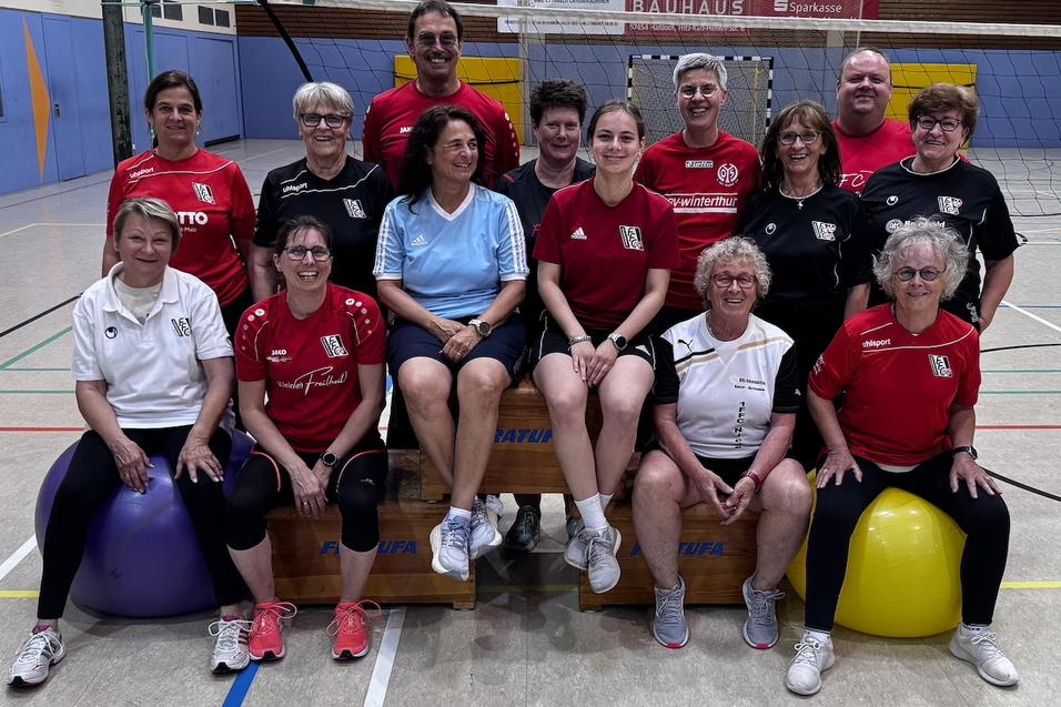 Gruppe von 15 Personen in Sportkleidung, l&auml;chelnd auf Boxen und B&auml;llen in einer Sporthalle. Volleyballnetz im Hintergrund.