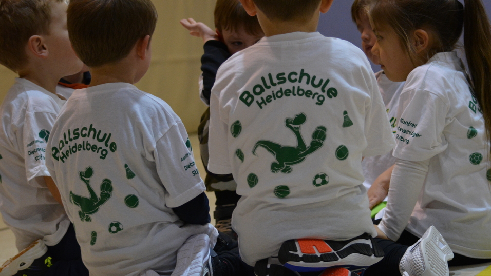 Sechs Kinder in wei&szlig;en T-Shirts mit Aufdruck sitzen im Kreis auf dem Boden und besprechen eine Sport&uuml;bung.