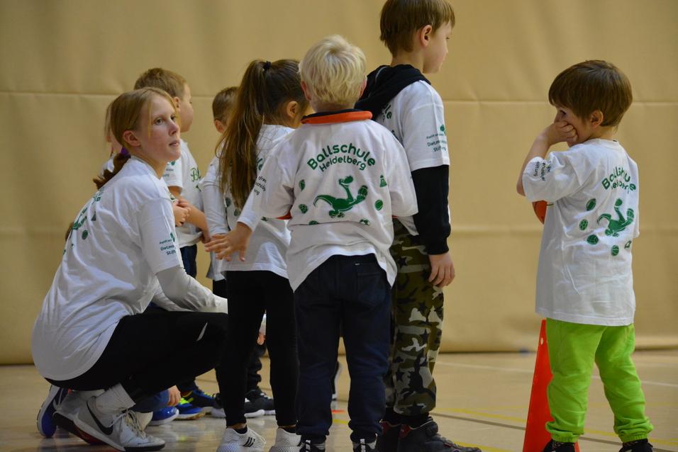 Eine Gruppe von Kindern in wei&szlig;en T-Shirts steht mit einer Betreuerin in einer Sporthalle.