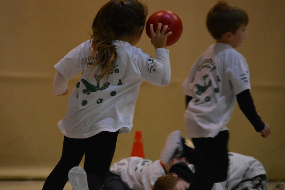 Ein Kind mit einem roten Ball in der Hand l&auml;uft &uuml;ber den Boden, w&auml;hrend andere Kinder in wei&szlig;en T-Shirts spielen.