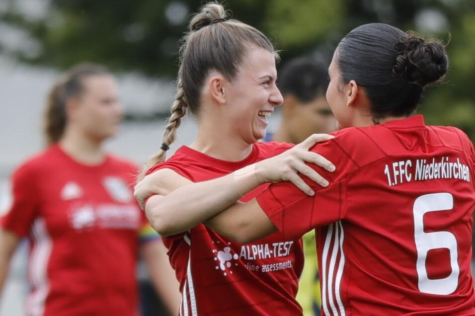 Zwei Fu&szlig;ballspielerinnen in roten Trikots umarmen sich l&auml;chelnd auf dem Spielfeld. Hintergrund unscharf.