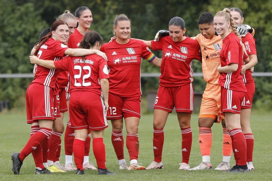 Mannschaftsbesprechung von Frauenfu&szlig;ballspielerinnen in roten Trikots auf einem Sportplatz, umarmt und l&auml;chelt.