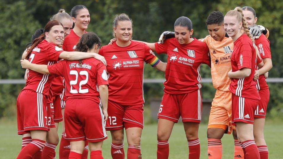 Mannschaftsbesprechung von Frauenfu&szlig;ballspielerinnen in roten Trikots auf einem Sportplatz, umarmt und l&auml;chelt.
