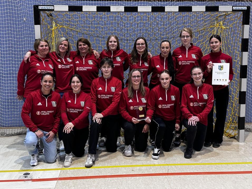 Mannschaftsbild einer Handballmannschaft in roten Trainingsanz&uuml;gen, gruppiert vor einem Tor mit Urkunde und Pokal.