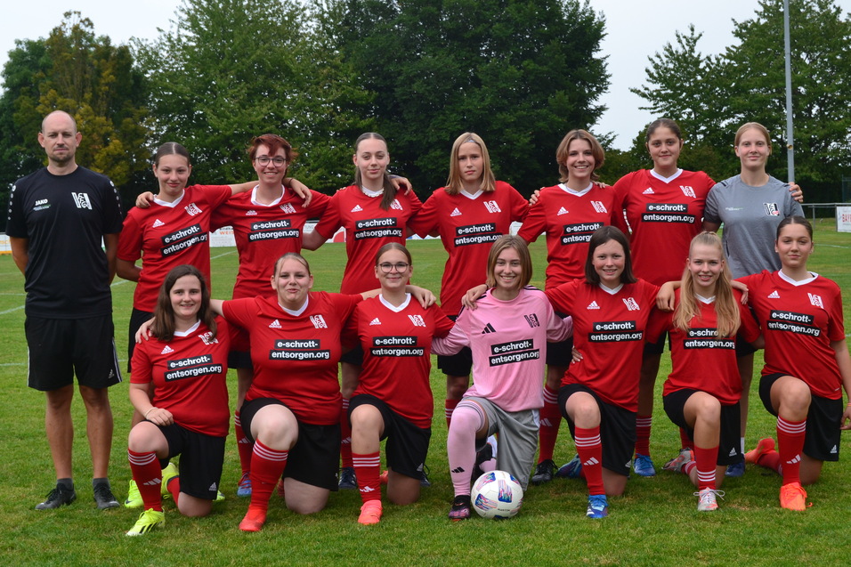Damenfu&szlig;ballmannschaft in roten Trikots posiert auf einem Sportplatz,Trainer steht links hinter den Spielerinnen.