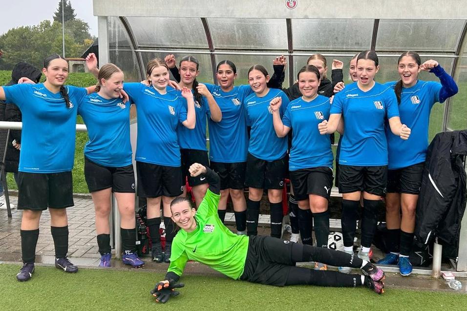 Gruppenszene einer Fu&szlig;ballmannschaft mit l&auml;chelnden Spielerinnen in blauen Trikots auf der Bank, eine Torh&uuml;terin liegt vorn.