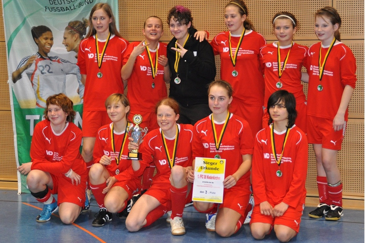 Gruppenshot von 12 jungen Fu&szlig;ballerinnen in roten Trikots mit Medaillen und Pokalen, l&auml;chelnd auf einem Hallenboden.