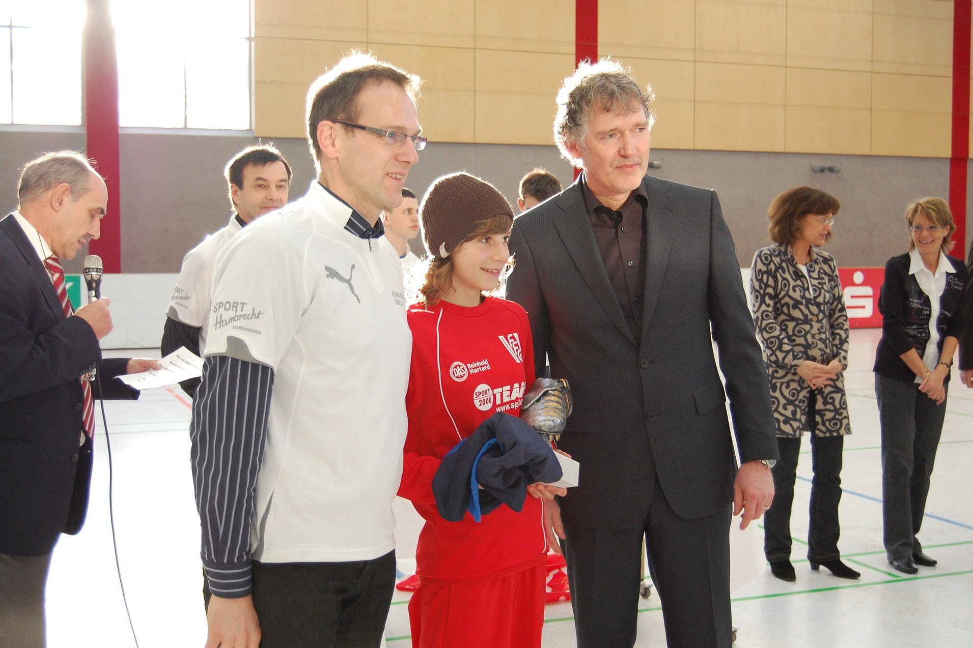 Zwei M&auml;nner und ein Jugendlicher in Sportkleidung posieren gemeinsam in einer Sporthalle f&uuml;r ein Foto.