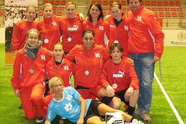 Gruppensfoto eines Frauenfu&szlig;ballteams mit Medaillen, stehend und sitzend auf einem Kunstrasenplatz.