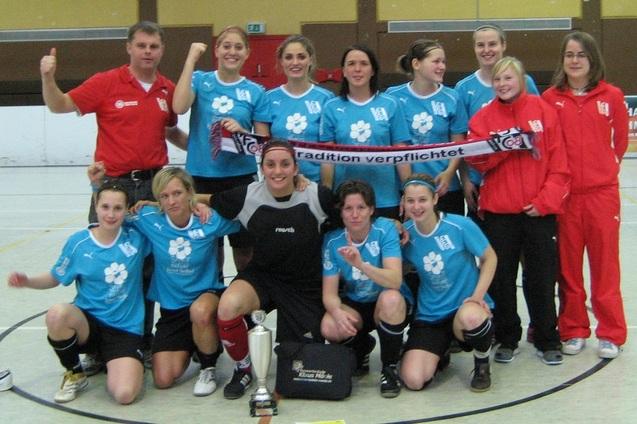 Frauenfu&szlig;ballmannschaft in blauen Trikots posiert mit Pokal und Banner in einer Sporthalle.