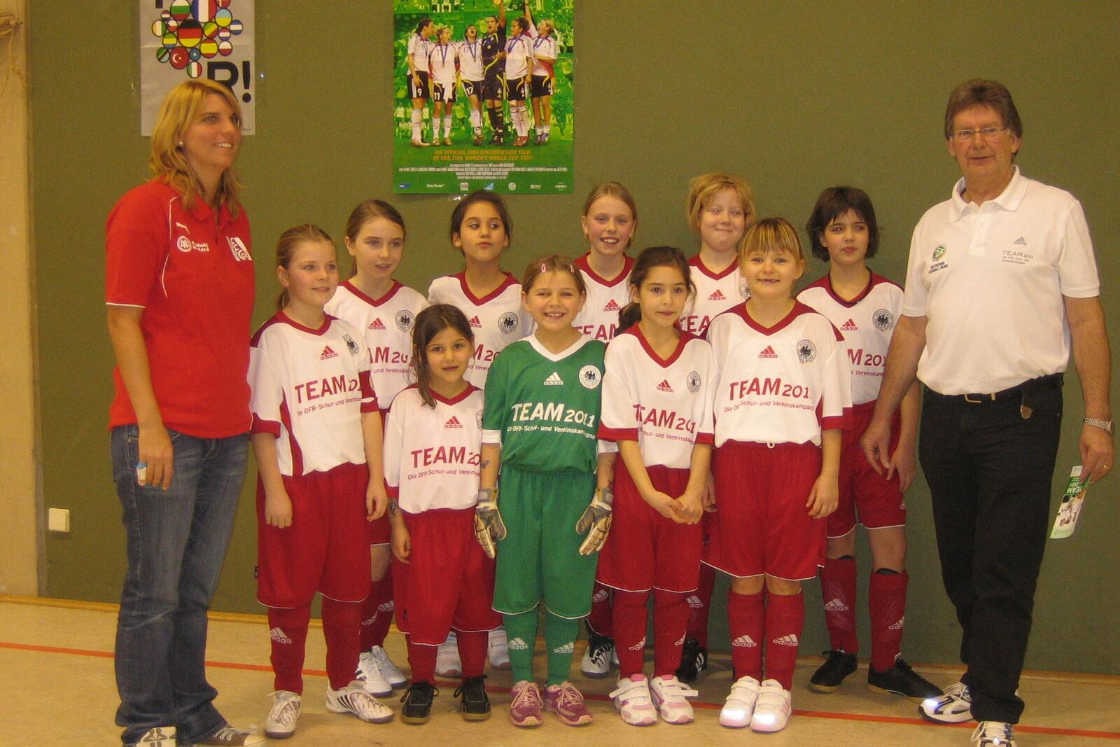 Mannschaftsaufstellung von jungen Fu&szlig;ballspielerinnen in roten Trikots, begleitet von Trainern, vor einem gr&uuml;nen Hintergrund.