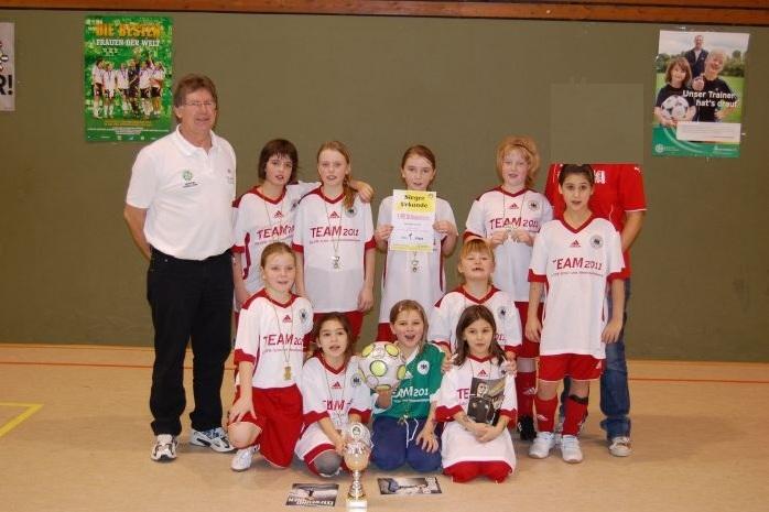 Gruppenfoto einer Mädchenfußballmannschaft mit Trainer, Pokalen, Urkunden und Ball in einer Sporthalle.