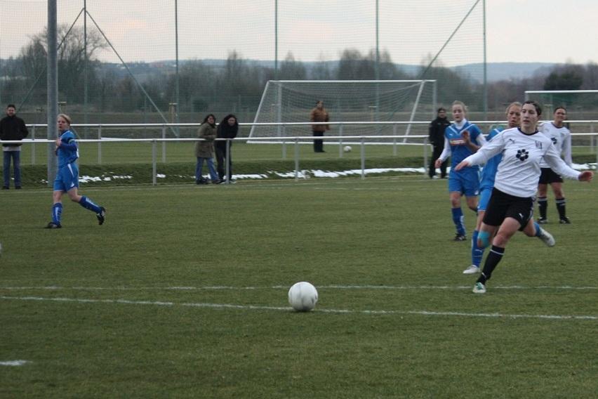 Spielszene auf einem Fu&szlig;ballplatz: Spielerinnen in blauen und wei&szlig;en Trikots verfolgen den Ball.
