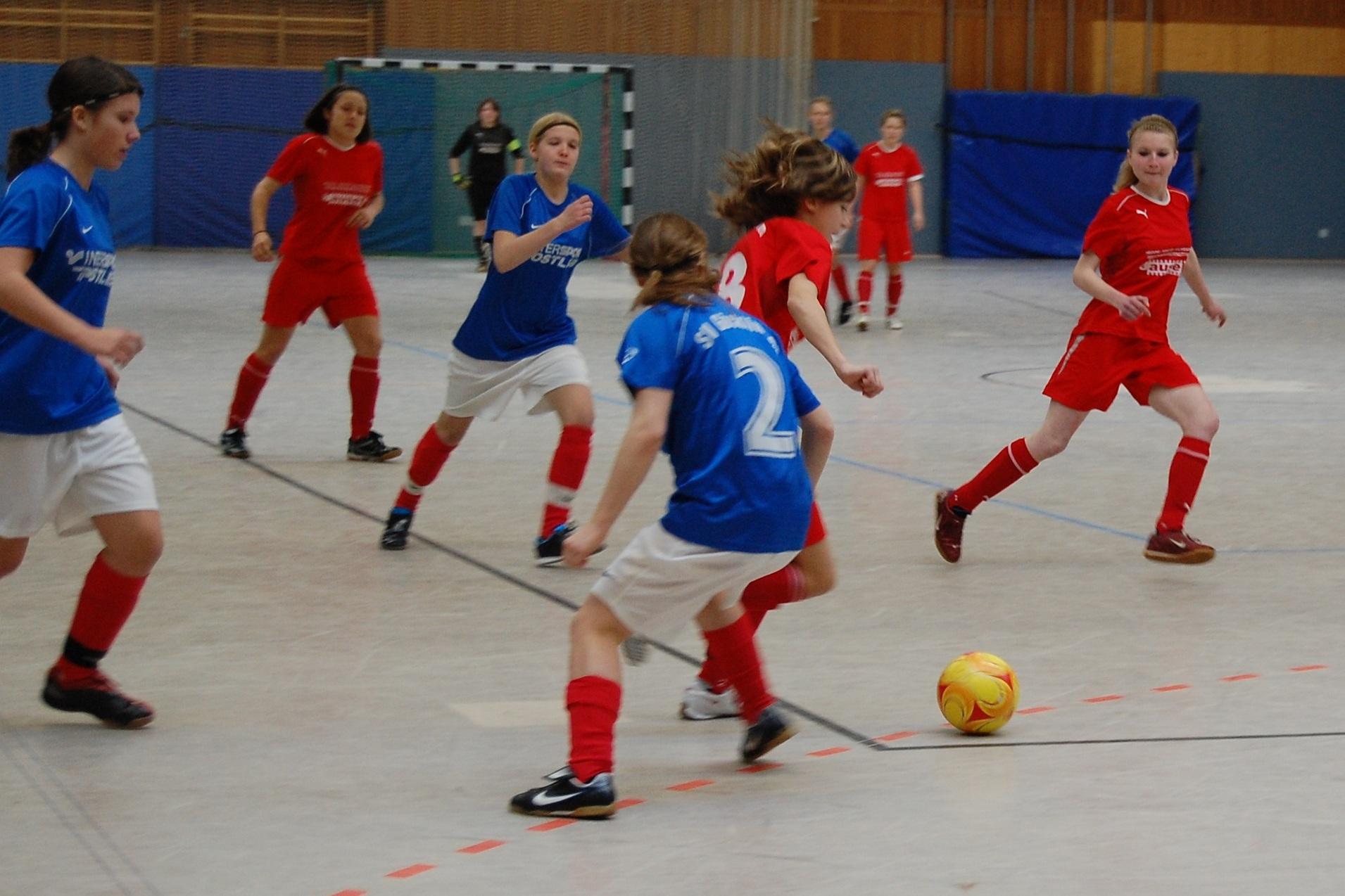 M&auml;dchenfu&szlig;ballspiel in einer Halle, zwei Teams in blauen und roten Trikots jagen nach einem gelben Ball.