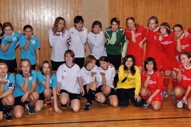 Gruppenfoto von zwei Fu&szlig;ballmannschaften mit M&auml;dchen in roten und blauen Trikots, l&auml;chelnd in einer Sporthalle.