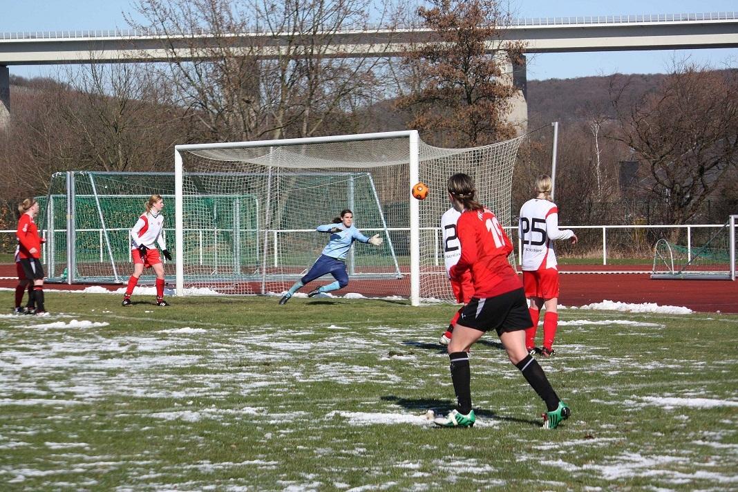 Spielszene eines Fußballspiels, Torwart springt nach Ball auf schneebedecktem Rasen, Spieler im roten Trikot im Hintergrund.