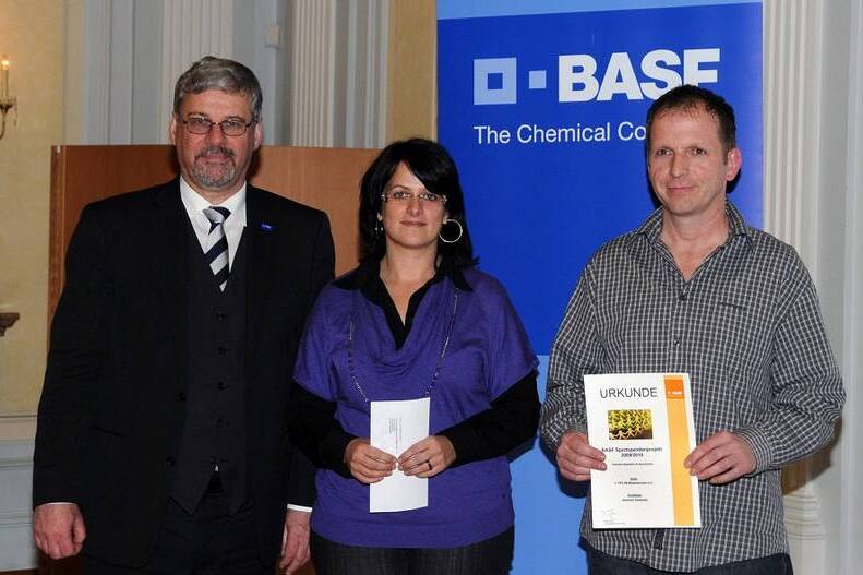 Drei Personen stehen vor einem BASE-Firmenschild, die Frau in der Mitte h&auml;lt ein Dokument, w&auml;hrend die M&auml;nner daneben stehen.