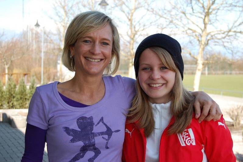 Zwei l&auml;chelnde Frauen stehen arm in arm auf einem Platz, im Hintergrund sind B&auml;ume und ein Sportfeld sichtbar.