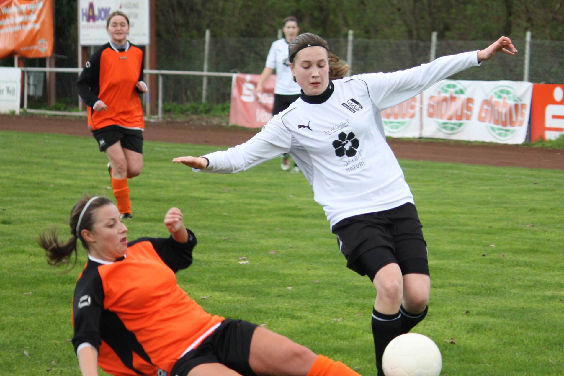 Zwei Fußballspielerinnen im Spiel: Eine in orange beim Rutschen, während die andere in weiß den Ball spielt.