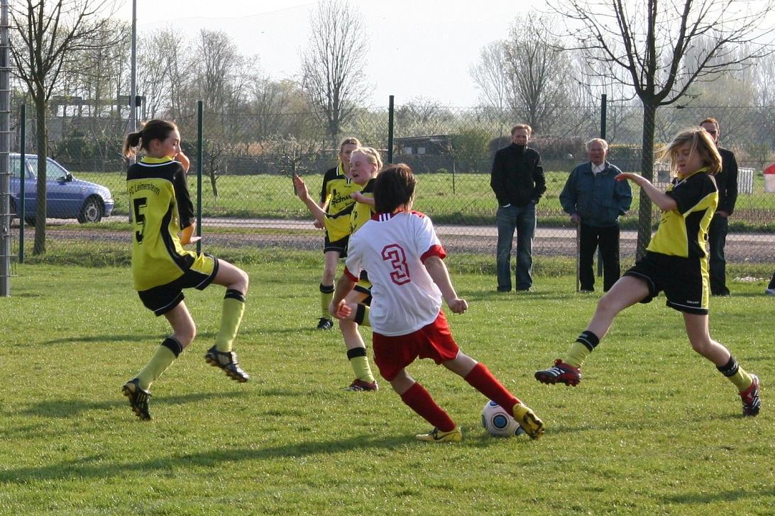 Zwei M&auml;dchen in gelben Trikots und ein Junge in rotem Trikot spielen Fu&szlig;ball auf einem Rasenplatz.